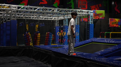 A young boy stands on the edge of a trampoline in an indoor adventure park. The park features obstacle courses and colorful padding.