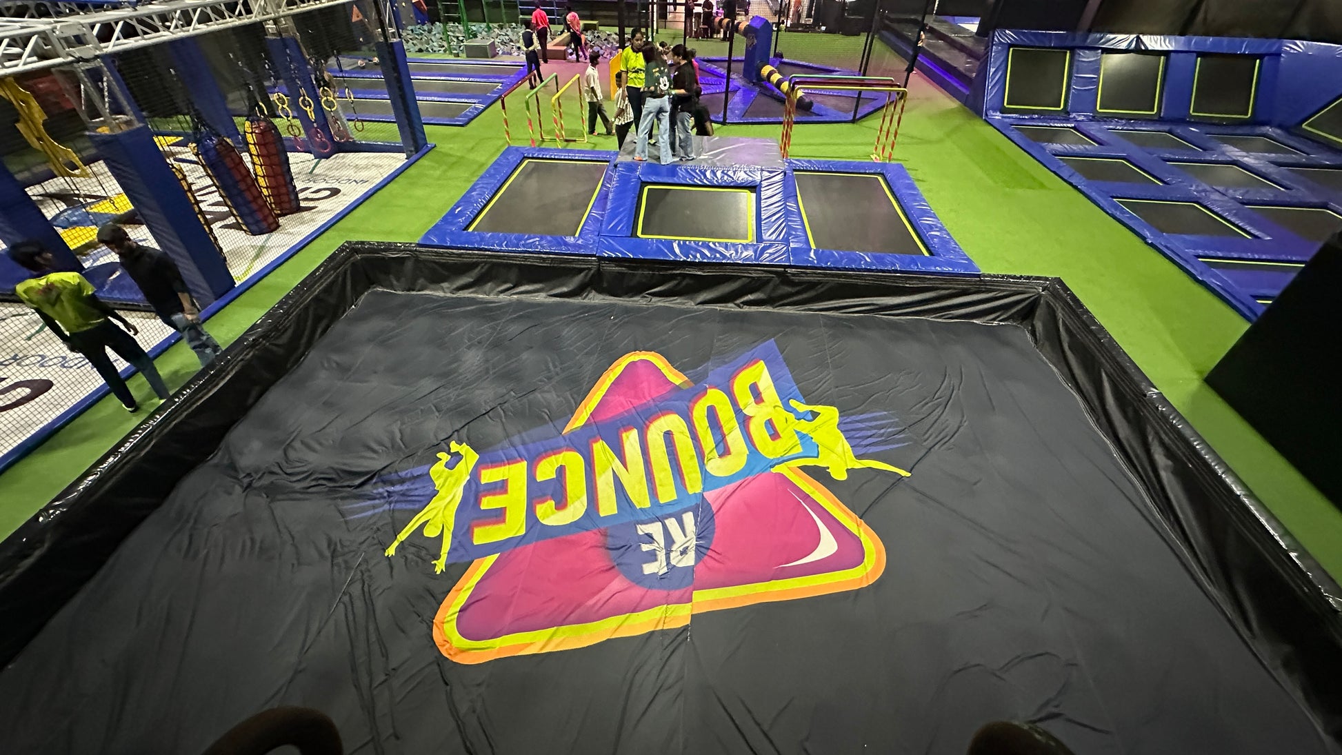 People jump on trampolines at an indoor trampoline park. A large "BOUNCE" logo is visible on a black mat.