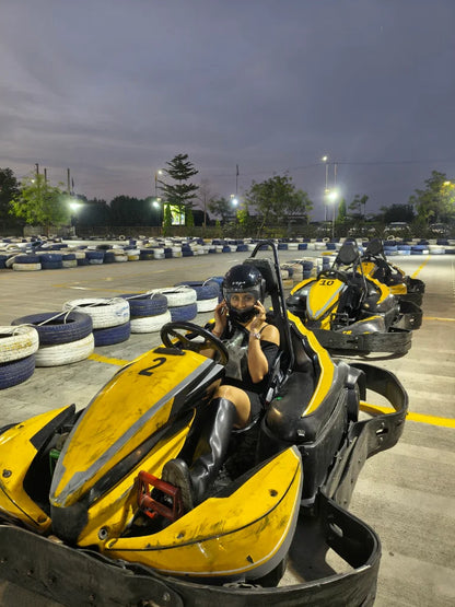A woman in a helmet and boots sits in a yellow go-kart on a track at dusk.