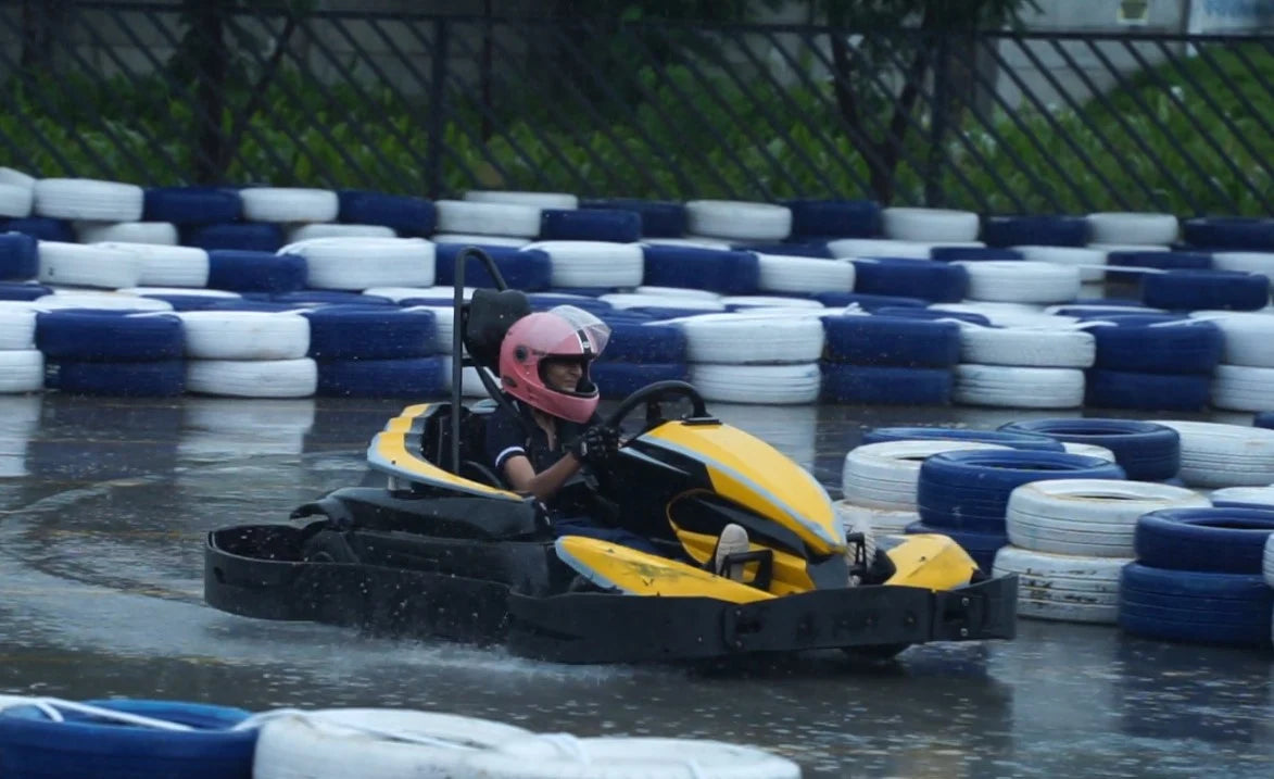 A young person in a pink helmet races a go-kart on a wet track, with tire barriers lining the course.