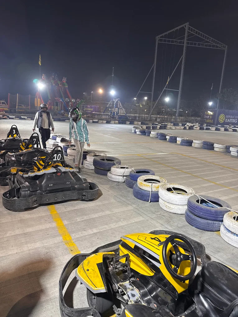 Go-karts are lined up on a track at night, with two young people in helmets standing nearby.