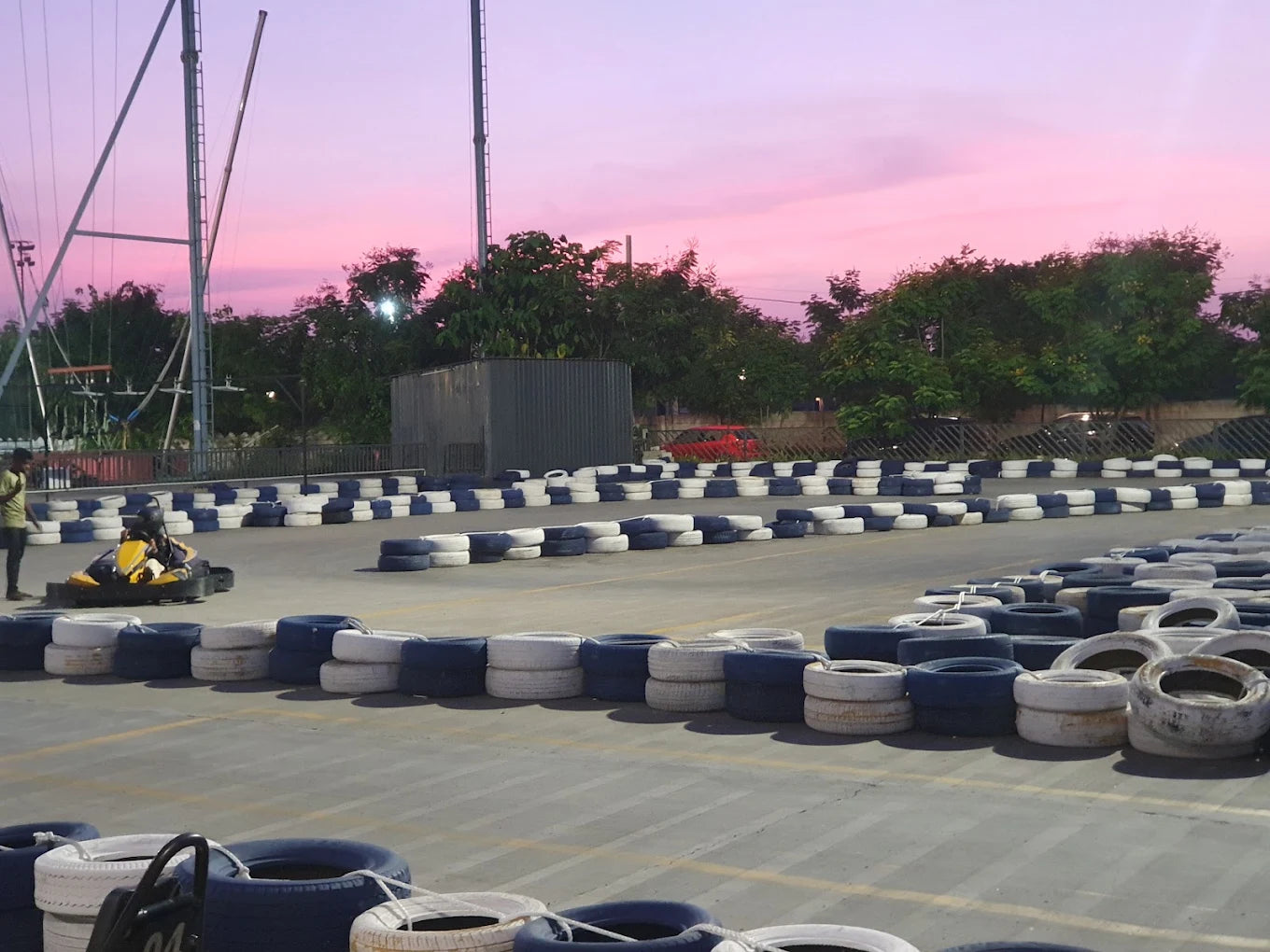 A go-kart track with a person driving a go-kart, bordered by tires, under a pink sunset sky.