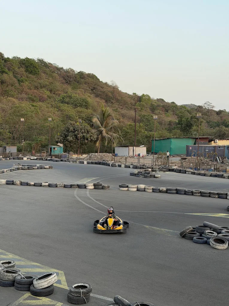 A child drives a go-kart on a race track with tire barriers, a wooded hill in the background.