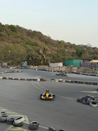 A child drives a go-kart on a race track with tire barriers, a wooded hill in the background.