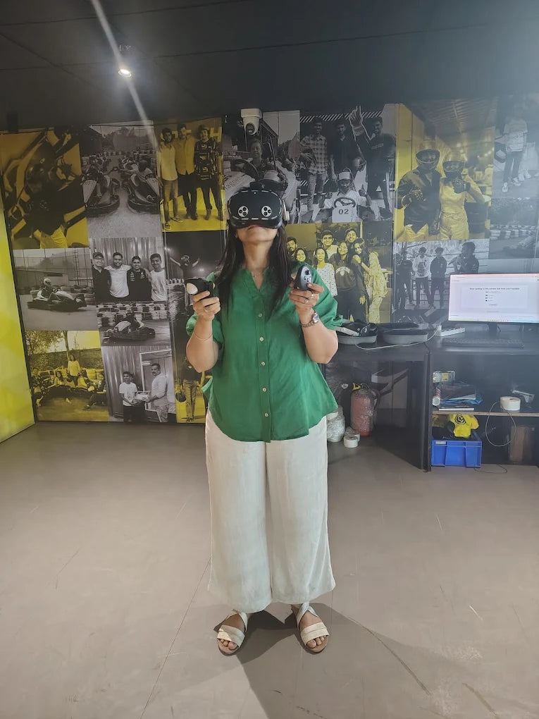 A young woman in a green shirt and white pants uses VR controllers and a headset in a room with a photo-covered wall.