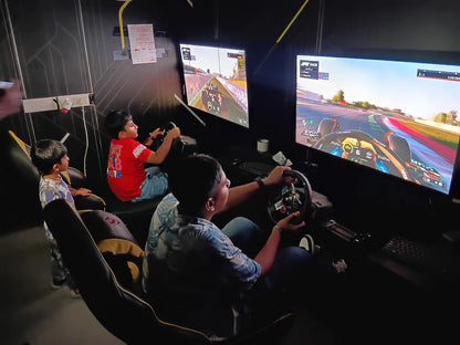 Children play a racing video game using steering wheels and monitors in a dark room.