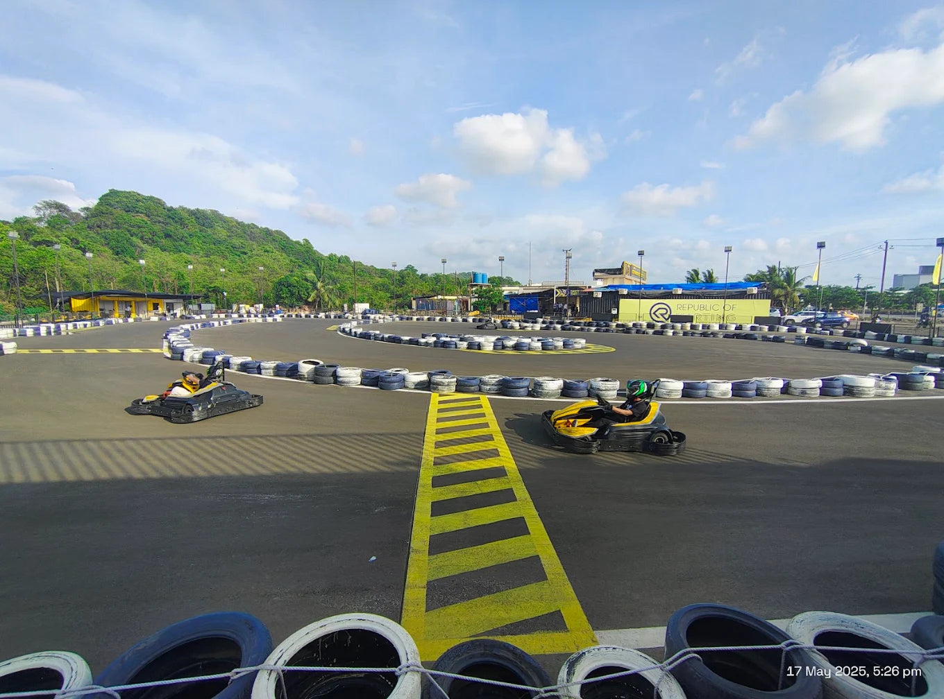 Two people race go-karts on a sunny outdoor track with lush green hills in the background.