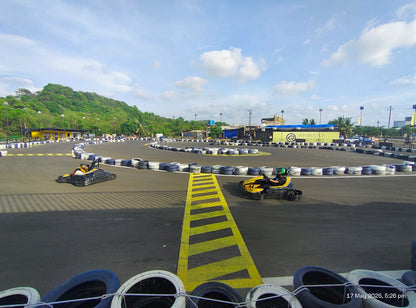 Two people race go-karts on a sunny outdoor track with lush green hills in the background.