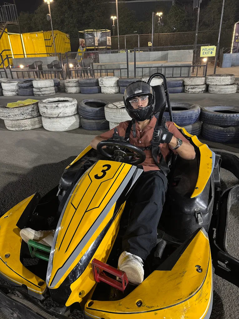 A young person in a go-kart helmet sits in a yellow go-kart at a track at night.