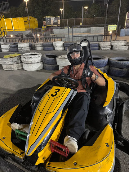A young person in a go-kart helmet sits in a yellow go-kart at a track at night.
