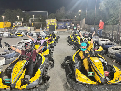 Adults in go-karts at a nighttime track, raising their hands in excitement.