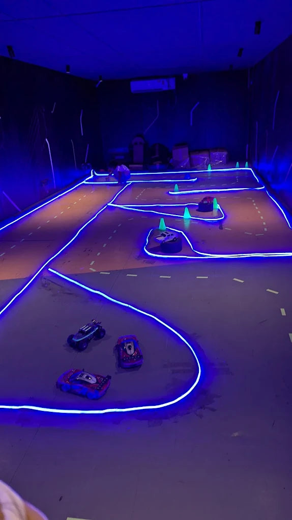 A group of children race remote-controlled cars on a neon-lit track in a dark room. The track is outlined with bright blue lights, and small green cones mark the course.