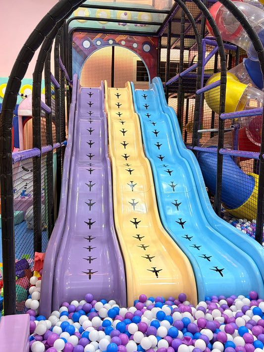 Three colorful slides in a children's play area, decorated with airplane decals, lead down to a ball pit.