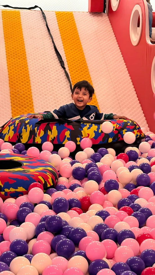 A young child laughs joyfully while sitting in a ball pit. The child is surrounded by colorful balls and a patterned inflatable.
