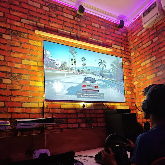 Person playing a racing game on a large screen in a room with brick walls.