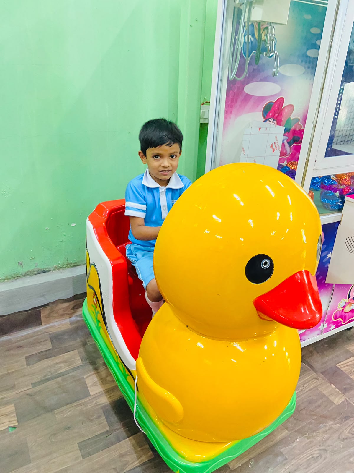 A young boy sits in a duck-shaped ride next to a claw machine.