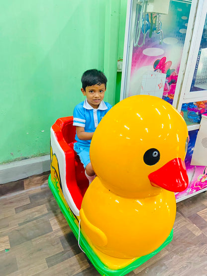 A young boy sits in a duck-shaped ride next to a claw machine.