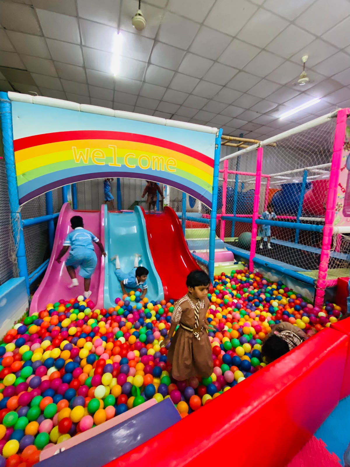 Children play in a colorful ball pit with slides under a rainbow "welcome" sign.