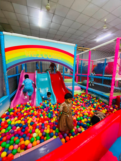 Children play in a colorful ball pit with slides under a rainbow "welcome" sign.