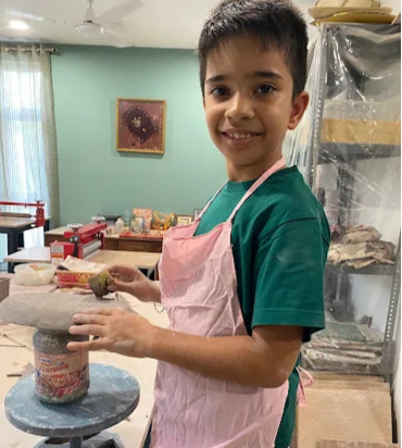 Child in a pottery studio with a pink apron and green shirt, smiling.
