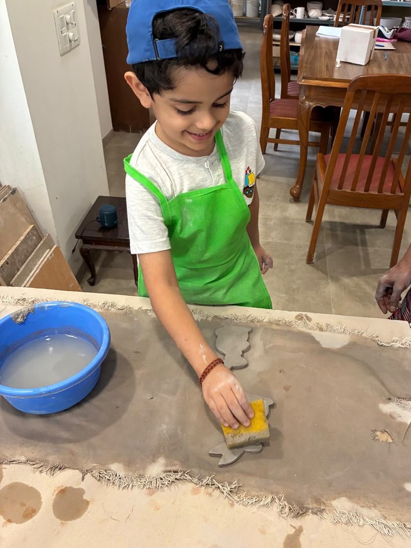 A young boy in a green apron works with clay, using a sponge to smooth a shape. He is at a table with a blue bucket of water.