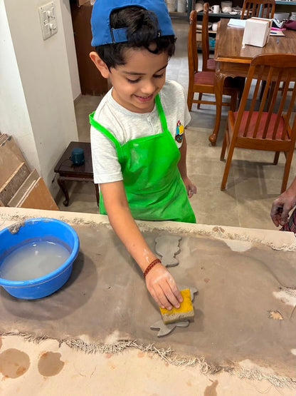 A young boy in a green apron works with clay, using a sponge to smooth a shape. He is at a table with a blue bucket of water.