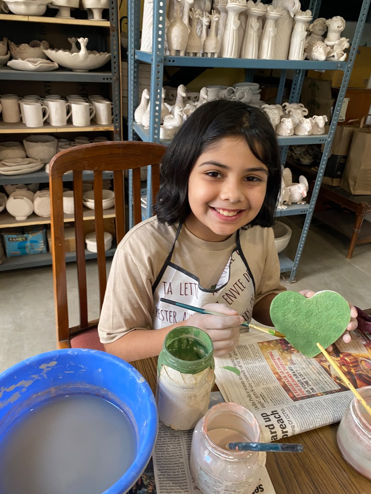 A young girl paints a green heart at a pottery studio, surrounded by shelves of ceramic pieces.