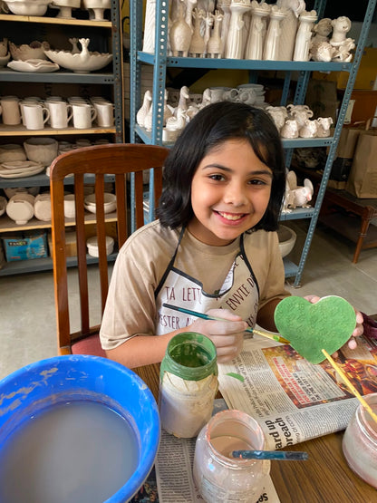 A young girl paints a green heart at a pottery studio, surrounded by shelves of ceramic pieces.