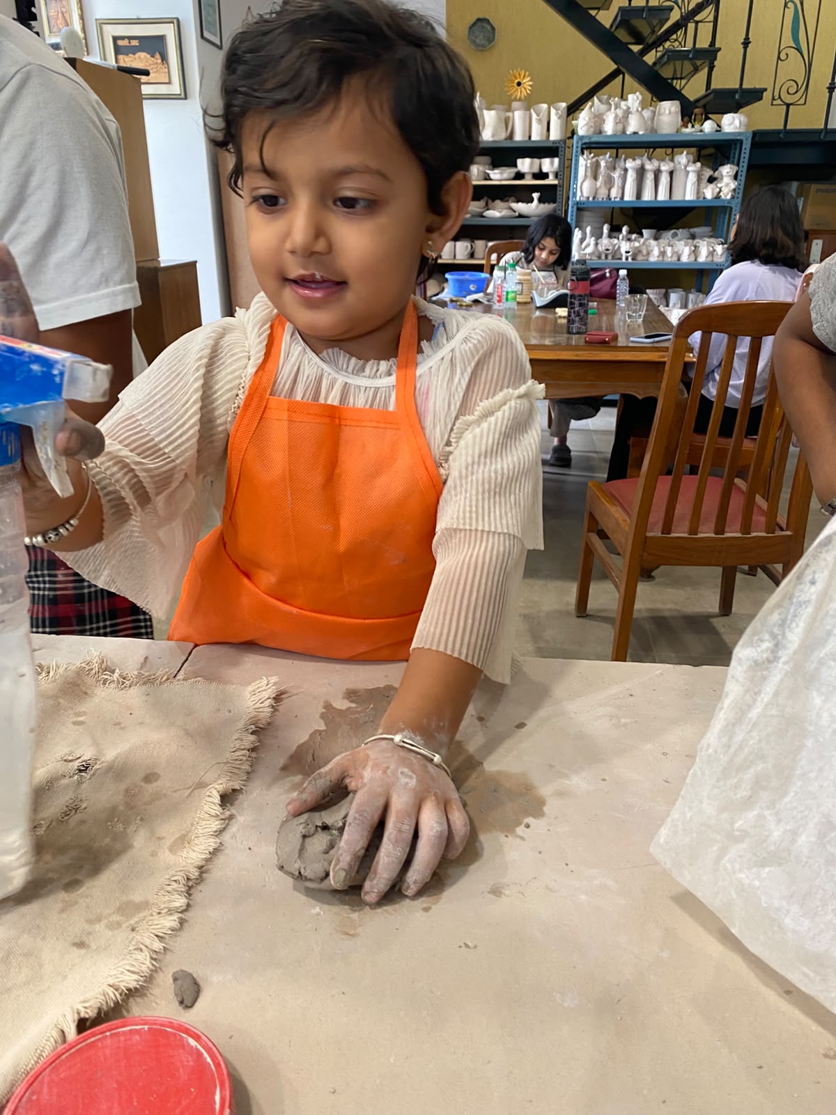 A young girl in an orange apron works with clay at a table, with shelves of pottery in the background.