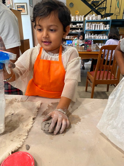 A young girl in an orange apron works with clay at a table, with shelves of pottery in the background.