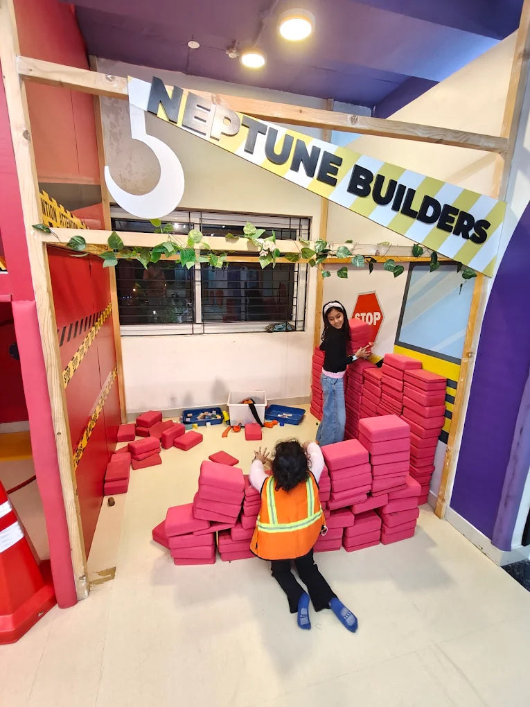Two young children play with large pink blocks in a building-themed play area. One child wears a construction vest.