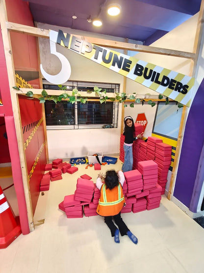 Two young children play with large pink blocks in a building-themed play area. One child wears a construction vest.