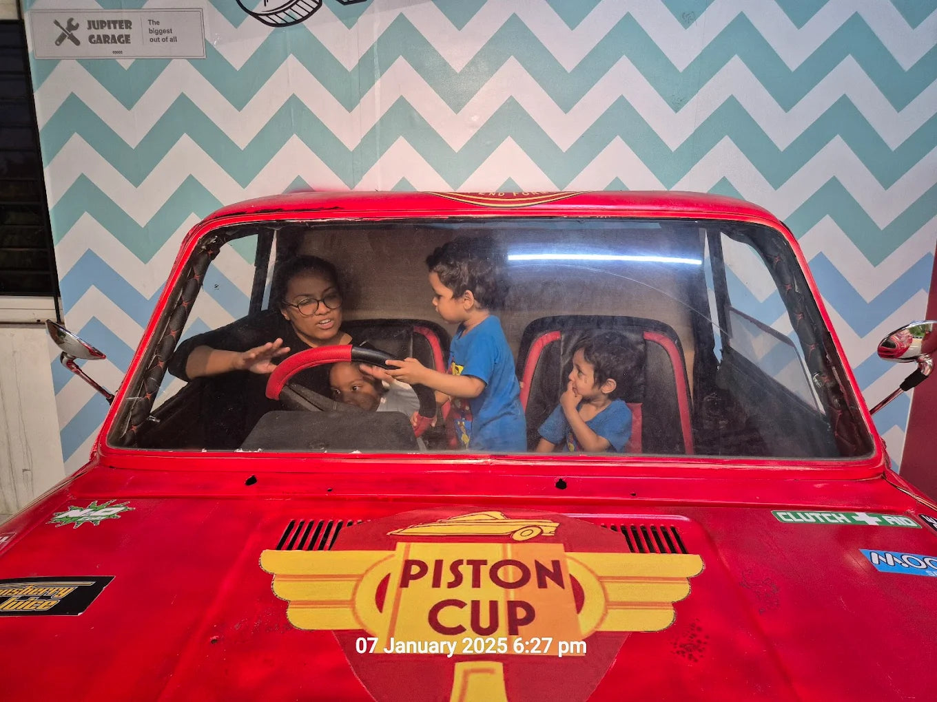 A woman and three young children sit in a red toy car with a "Piston Cup" decal.