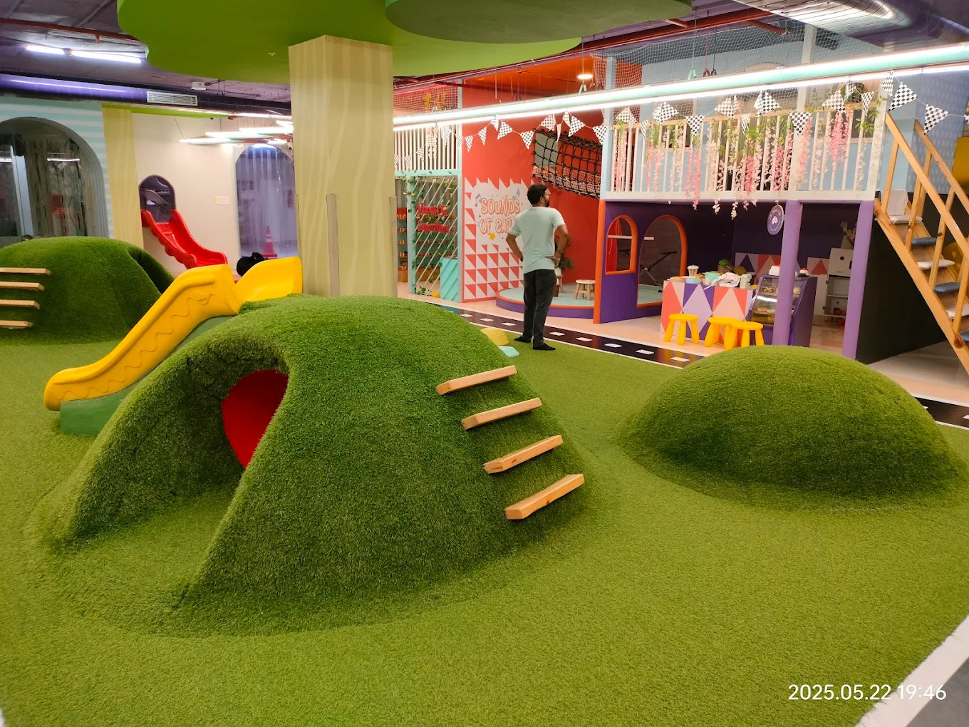 A man stands in a colorful indoor playground with slides and soft green hills.