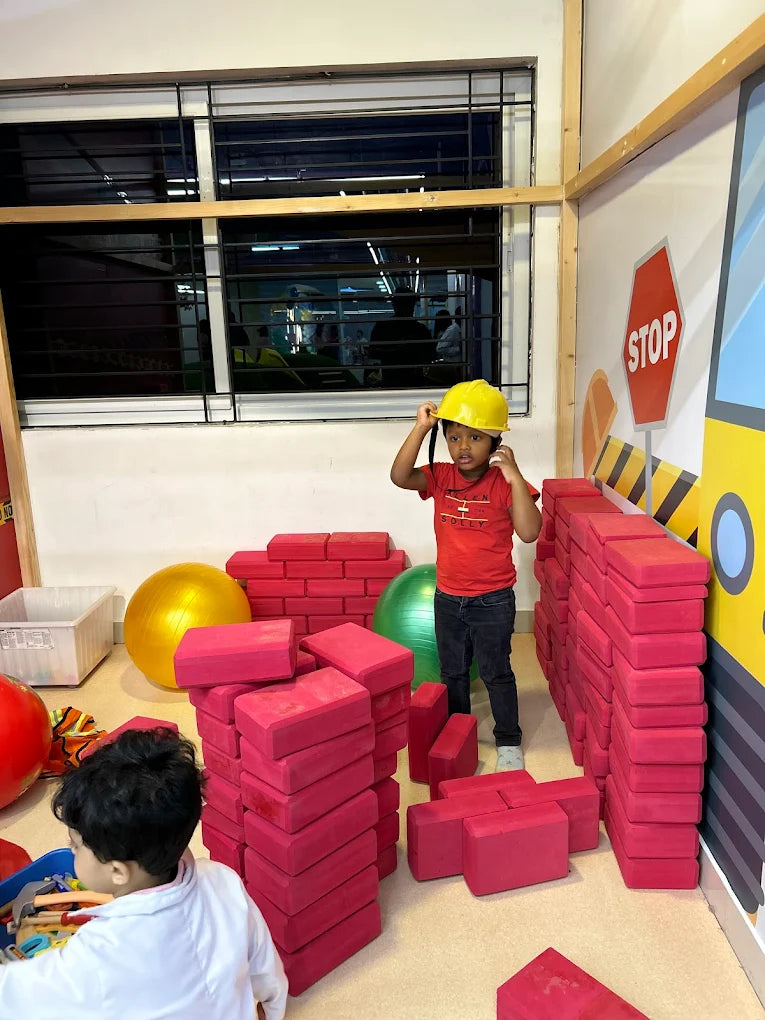 A young boy in a yellow hard hat plays with large pink building blocks in a room with a stop sign decoration.