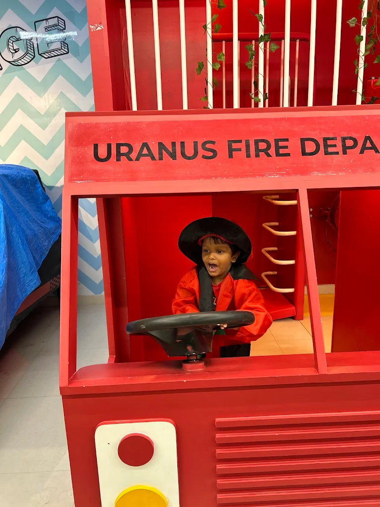 A young child in a firefighter costume plays at the wheel of a red fire truck. The truck has "URANUS FIRE DEPA" written on it.