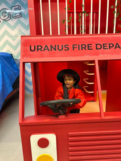 A young child in a firefighter costume plays at the wheel of a red fire truck. The truck has "URANUS FIRE DEPA" written on it.