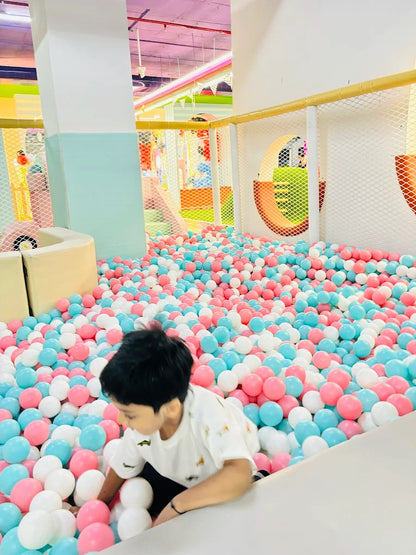 A young boy plays in a ball pit filled with pink, blue, and white balls.