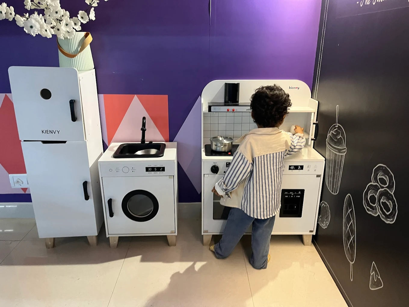 A young child plays with a toy kitchen set in a room with purple walls and chalk drawings.
