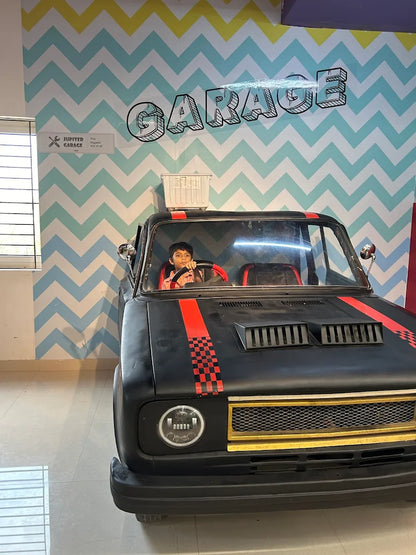 A child sits in the driver's seat of a black toy car with red racing stripes, in front of a chevron-patterned wall that says "GARAGE".