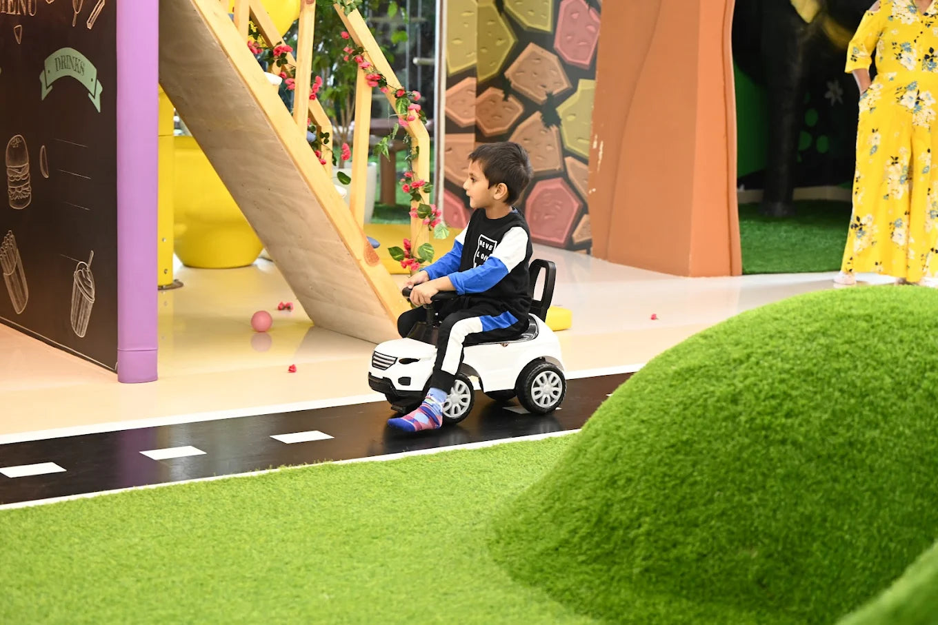 A young boy rides a toy car on a road in a colorful play area. He is looking to the side, wearing a black and blue shirt and striped pants.