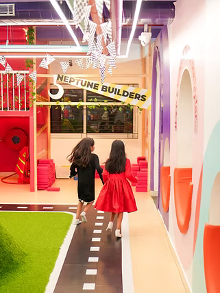 Two young girls in dresses run down a colorful indoor play area with a road design on the floor.