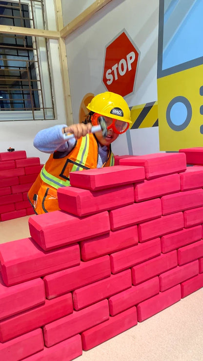 A child in a hard hat and vest pretends to build a wall with pink blocks.