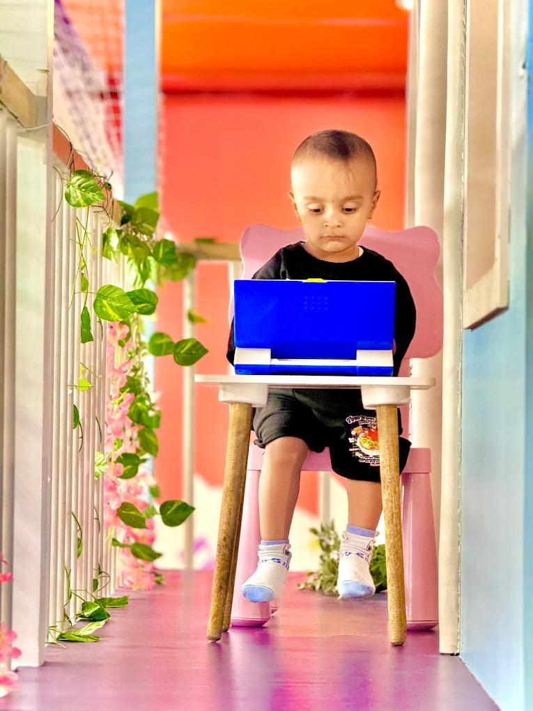 A young child sits at a small table, looking at a blue toy laptop in a colorful, playful room.
