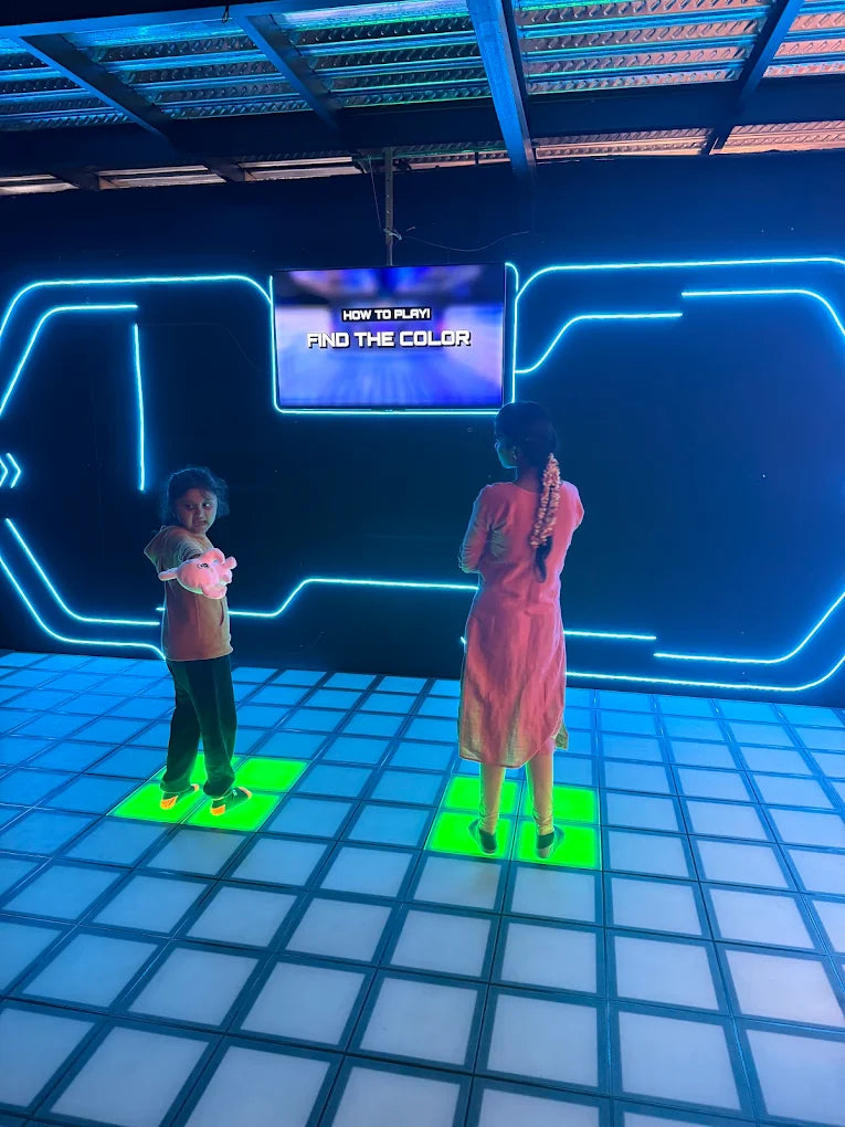 Two children stand on a glowing floor in front of a screen that says "FIND THE COLOR."