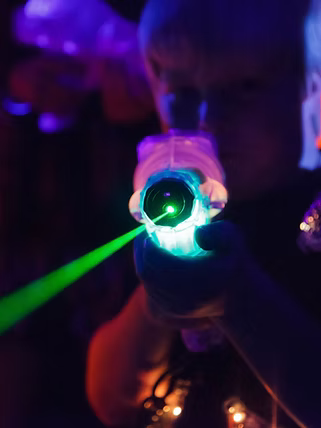 A child aims a glowing laser tag gun, its green beam cutting through the dark, neon-lit arena.