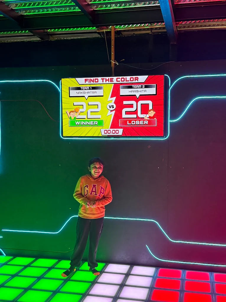 A young child stands on a colorful, illuminated floor in front of a game screen displaying "FIND THE COLOR" with scores for two teams.