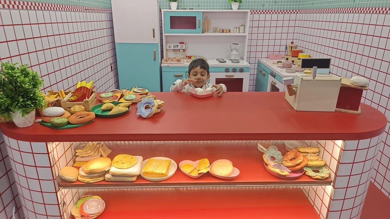 A young child plays with toy food in a play kitchen. The child is behind a red counter filled with toy food items.