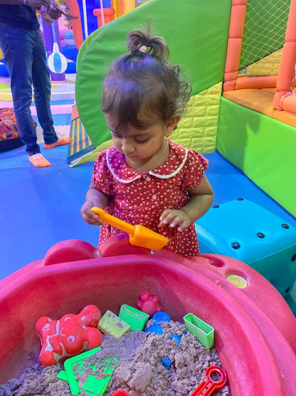 A toddler plays with kinetic sand in a red sandbox at an indoor playground.