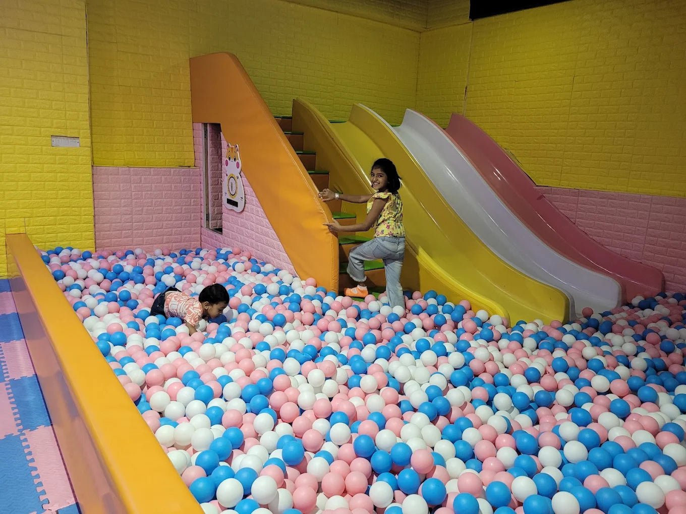 Two young children play in a ball pit with slides in the background.